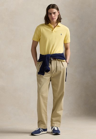 Mladić s smeđom kosom do ramena, nosi žuti polo, bež hlače, mornarsko plavi džemper vezan oko struka i plave tenisice.