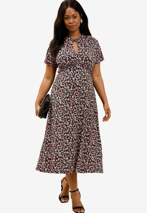Schwarzes Blumenkleid mit kurzen Ärmeln, das mit rosa und weißen Blumen verziert ist, mit einem Schlüsselausschnitt und einem mittellangen Rock. Trägt schwarze Sandalen.