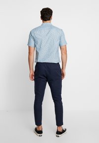 Man som står med ryggen mot kameran, iförd en ljusblå kortärmad skjorta med små mönster, marinblå byxor och svarta låga sneakers.