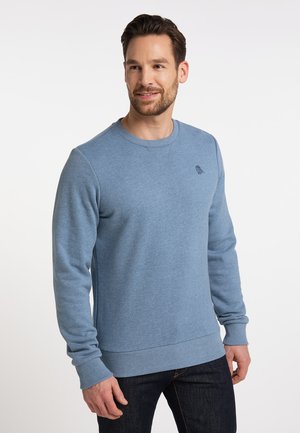 Uomo con capelli corti castani e barba, che indossa un maglione celeste a maniche lunghe e jeans scuri, guarda leggermente di lato sorridendo.