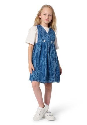 Blauwe denim jurk met bladpatroon, voorzien van een uitlopende rok, verstelbare bandjes en twee knopen aan de voorkant, gecombineerd met witte sneakers met klittenbandsluiting.