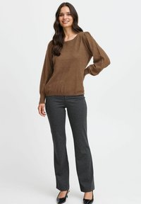 Brauner strukturiierter Pullover mit langen puffärmeln, kombiniert mit dunkelgrauen engen Hosen und schwarzen Absatzschuhen. Einfarbiger Hintergrund.