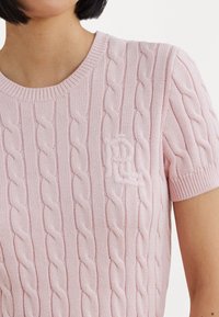 Růžový pletený svetr s copánkovým vzorem, krátkými rukávy a žebrovaným výstřihem. Na hrudi je vyšívané logo.