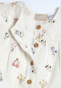 Chemisier brodé floral en coton crème, avec des boutons en bois, des manches courtes à volants et des détails en œillets. Motifs de fleurs colorées sur l'ensemble.