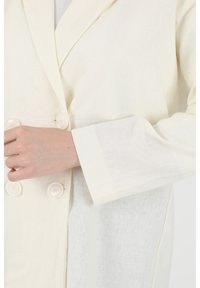 Light cream linen blazer with a double-breasted design, large buttons, and a relaxed fit. Features a notched collar and wide sleeves.