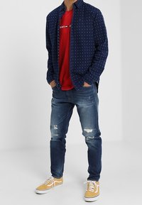 Námořnická puntíkovaná košile na knoflíky přes červené tričko, doplněná roztrhanými modrými džínami a žlutými teniskami, představující ležérní styl.