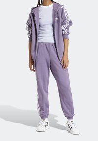 Lavender tracksuit with white Adidas stripes, featuring a zip-up hooded jacket and elastic waistband pants. Black and white sneakers complete the look.