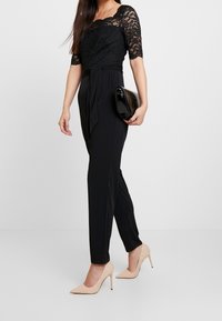 Top negro de encaje con mangas de tres cuartos, combinado con pantalones negros de cintura alta y tacones nude. Bolso de mano en material negro brillante.
