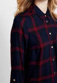 Marineblauwe en bordeauxrode geruite overhemd met kraag en gouden knopen, voorzien van lange mouwen en een patchzak op de borst.