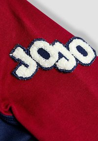 Material roșu cu logo-ul brodat "yojo" în textură albă asemănătoare blănii, conturat în nuanță de marină. Prim-plan care arată detalii ale materialului și designului.