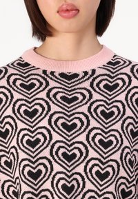 Maglione lavorato a maglia con scollatura rotonda a coste rosa e un motivo di cuori neri di diverse dimensioni su uno sfondo rosa pastello.