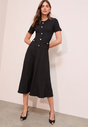 Donna in piedi contro il muro con un vestito midi nero testurizzato, maniche corte, bottoni sul davanti, tasche laterali e tacchi neri con punta appuntita.