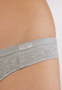 Szary bawełniany thong z miękkim elastycznym paskiem, na którym znajduje się metka z napisem "Intimissimi Natural." Gładka tekstura, zbliżenie na biodro.