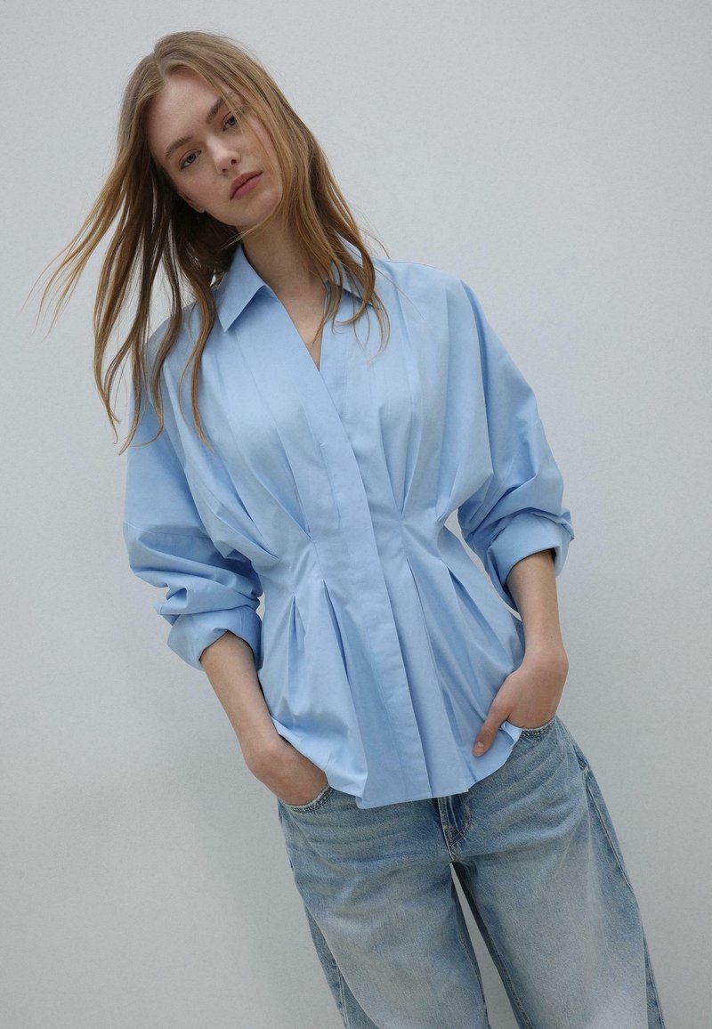 Jeune femme aux longs cheveux portant un chemisier plissé bleu clair et un jean bleu clair, debout les mains dans les poches devant un fond uni.