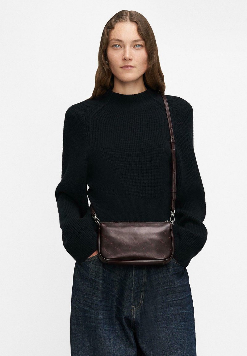 Kobieta z długimi brązowymi włosami nosi czarny sweter z dzianiny, ciemne dżinsy o szerokich nogawkach oraz małą, ciemno-brązową torbę na ramię.