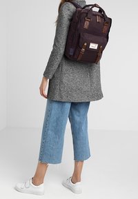 Sac à dos en tissu violet foncé avec accents en cuir marron, plusieurs compartiments, forme rectangulaire, porté sur le dos d'une personne, assorti à une tenue décontractée.