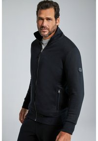 Chaqueta negra con cremallera y cuello alto, hecha de un tejido suave. Cuenta con bolsillos laterales y puños acanalados, diseñada para brindar comodidad y calidez.