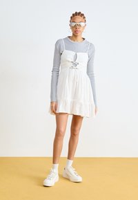 Vestido blanco con un dobladillo con volantes y diseño de abertura, superpuesto sobre una blusa de manga larga gris claro. Combinado con zapatillas blancas y gafas de sol.