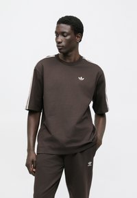 Brown cotton t-shirt with short sleeves, crew neck, and white Adidas logo. Features white stripes on shoulders, paired with matching pants.