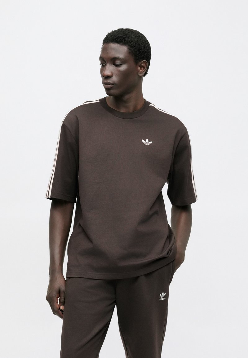 Brown cotton t-shirt with short sleeves, crew neck, and white Adidas logo. Features white stripes on shoulders, paired with matching pants.