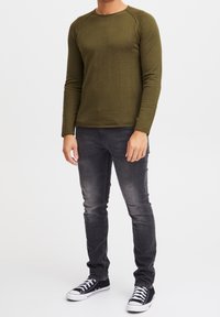 Olijfgroene lange mouwen shirt met een ronde hals en een gestructureerd patroon, gecombineerd met vervaagde zwarte strakke jeans en zwarte sneakers met witte zolen.
