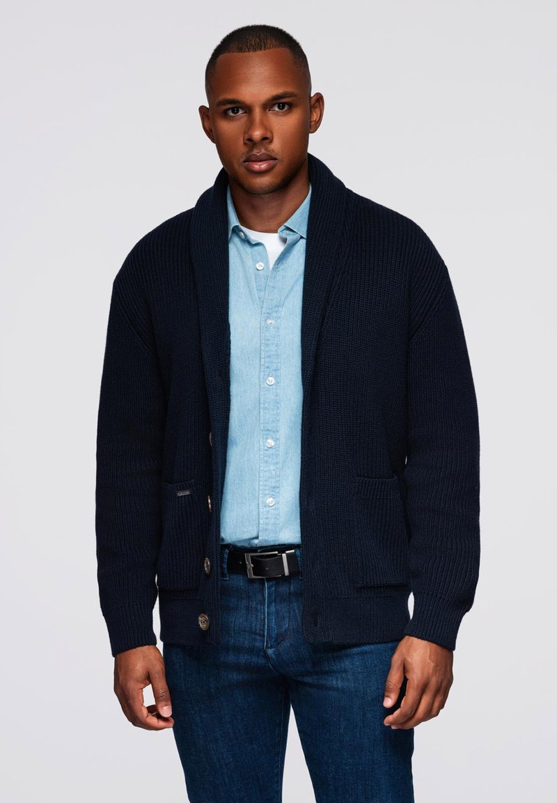 Cardigan lavorato a maglia blu navy con colletto a scialle, caratterizzato da chiusure con bottoni e tasche frontali, indossato sopra una camicia di denim blu chiaro e jeans scuri.