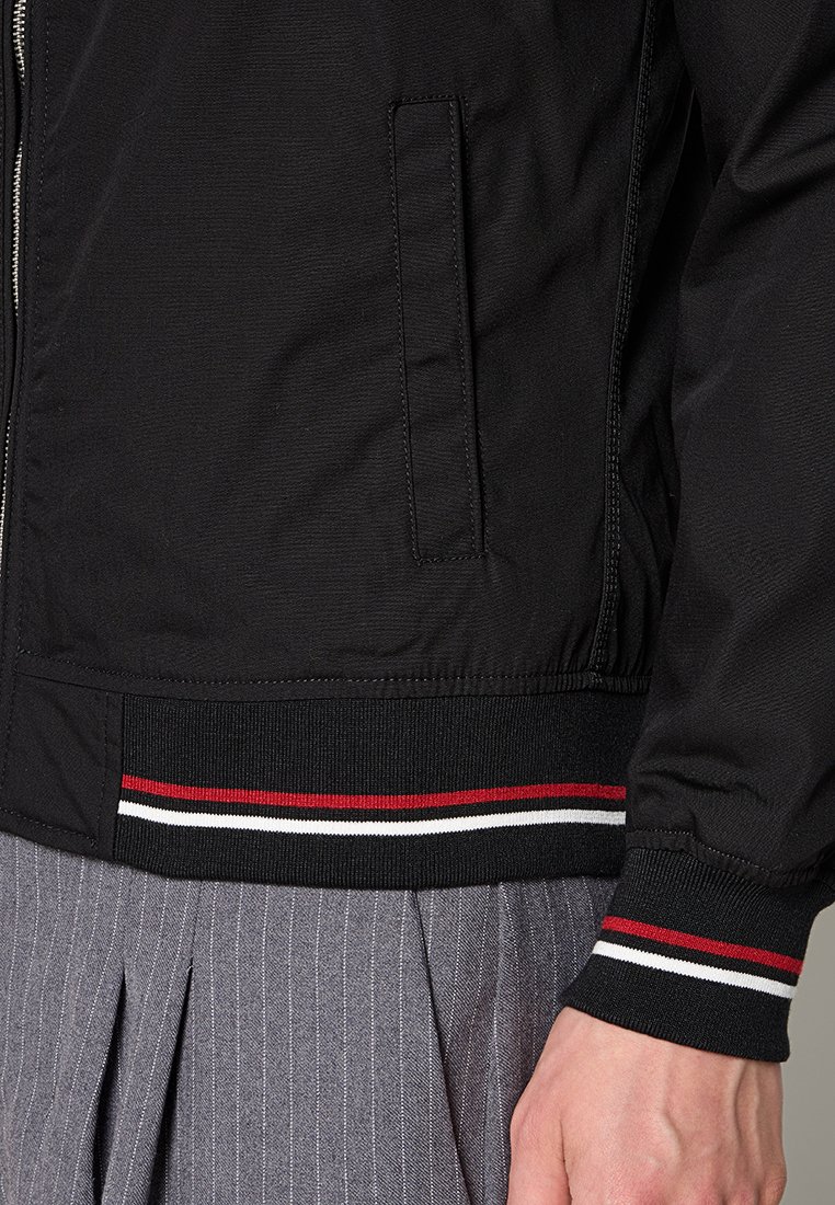 Veste bomber noire avec fermeture éclair, featuring un ourlet et des poignets côtelés avec des rayures rouge, blanche et noire ; texture lisse, sans matériel visible.