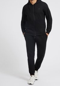Felpa nera con zip, cappuccio con coulisse e logo sul petto. Abbinata a pantaloni jogger neri e scarpe da ginnastica bianche.