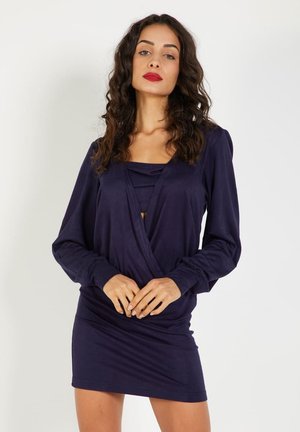 Mujer con cabello rizado vestida con un vestido mini de manga larga en azul marino, de pie contra un fondo blanco liso.