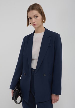 Blazer bleu marine croisé en tissu lisse, avec des revers crantés et deux boutons, accompagné d'un pull léger à côtes.