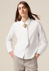 Chemise blanche à boutons avec un col pointu, dotée d'un détail de broche en or. Les manches longues ont des poignets arrondis ; le tissu semble lisse et léger.