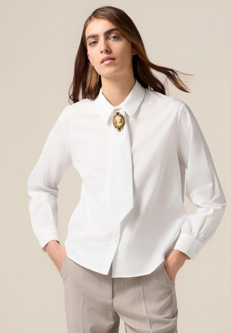 Chemise blanche à boutons avec un col pointu, dotée d'un détail de broche en or. Les manches longues ont des poignets arrondis ; le tissu semble lisse et léger.