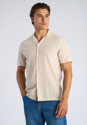 Camicia beige a maniche corte con motivo a coste strutturato e colletto a revers, chiusura frontale con bottoni, indossata con jeans di denim.