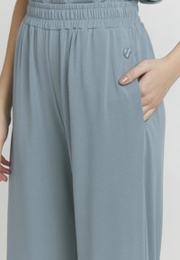 Mano che riposa nella tasca di pantaloni larghi e azzurri con vita elastica e piccolo distintivo rotondo in metallo sul lato destro.