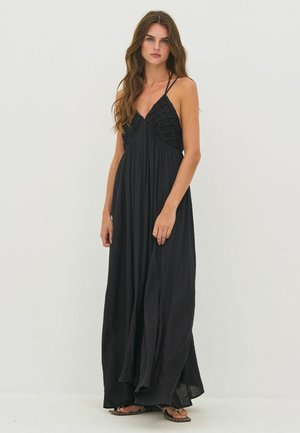 Pitkä- ja aaltohiuksinen nainen, jolla on musta hiaton maksi-mekko, jossa on kudottu yläosa ja ohuet olkaimet, seisomassa yksinkertaista vaaleaa taustaa vasten.