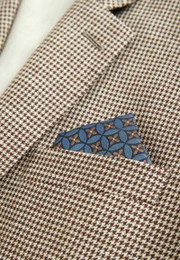 Hnedo-biely sakový vzor houndstooth s modrým geometrickým vzorovaným vreckovkou v náprsnom vrecku.