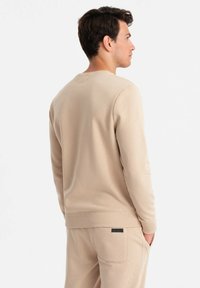 Beige Pullover mit geripptem Halsausschnitt und Bündchen, aus strukturiertem Stoff, getragen mit passenden beigen Hosen. Die Rückansicht zeigt ein minimalistisches Design.