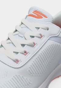 Λευκό αθλητικό παπούτσι με υφαντό επάνω μέρος με υφή, λευκά κορδόνια, ροζ θηλιές κορδονιών και πορτοκαλί λογότυπο στη γλώσσα.