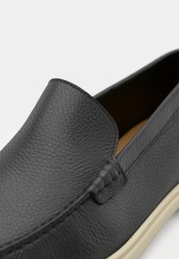 Black leather slip-on shoe with textured finish, rounded toe, and subtle stitching detail. Cream-colored sole adds contrast to design.