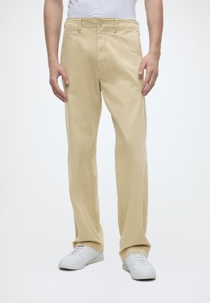 Homme portant un pantalon beige droit et des baskets blanches, debout les mains détendues le long du corps devant un fond uni.