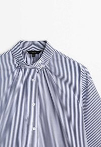 Chemise boutonnée à rayures verticales bleues et blanches avec un col haut orné de deux boutons sur le côté, posée à plat sur une surface blanche.