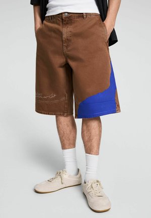 Mann trägt braune knielange Shorts mit einem blauen Fleck am rechten Bein, weiße Socken und beige Turnschuhe, steht mit den Händen in den Taschen.