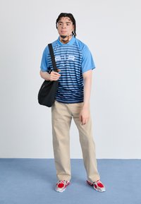 Light blue and navy striped athletic shirt with “AIR” logo, beige pants, and red and silver running shoes. Black shoulder bag.