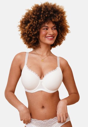 Woman with curly hair smiling, wearing a white padded bra with lace trim and matching lace underwear, standing against a plain background.