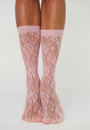 Beine mit kniehohen, rosa Spitzenstrümpfen mit Blumen- und Netzdetails vor hellem, einfarbigem Hintergrund.