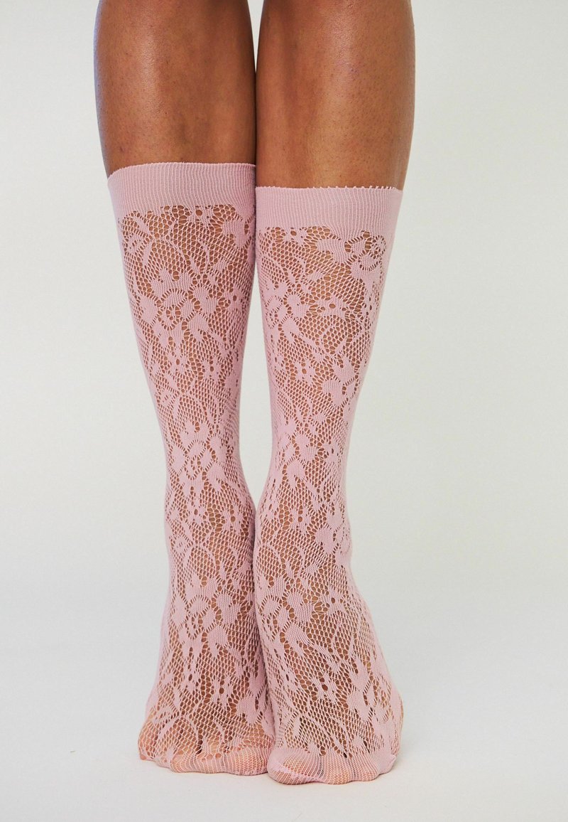 Jambes portant des chaussettes hautes roses à motif de dentelle avec des détails floraux et en résille sur un fond clair uni.