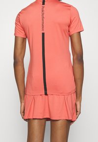 Top sportivo color pesca con maniche corte e gonna plissettata. Presenta una striscia verticale nera con il testo "Peak Performance" sul retro.