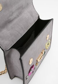 Borsa in suede grigia con dettagli neri, dotata di chiusura in oro e ornamenti decorativi tra cui paillettes e una toppa floreale.