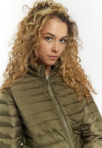 Khaki pufjakke med lynlås foran, der har en høj krave og horisontale syninger. Modellen har krøllet hår og står med ansigtet vendt fremad.