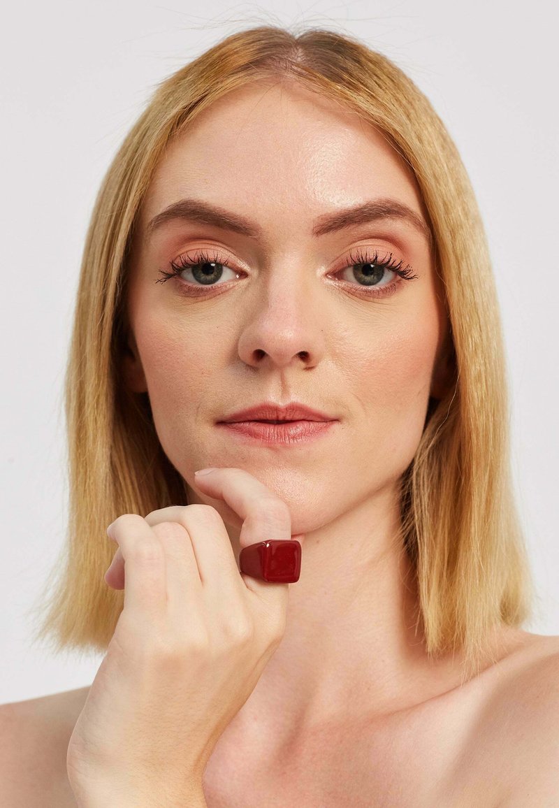 Crvena smolasta prsten s glatkom, sjajnom površinom i kvadratnim oblikom, nosi se na kažiprstu, u kombinaciji s neutralnim tonovima kože.